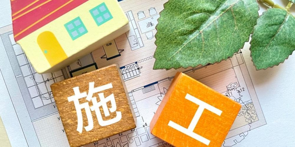 間取り図の上に置かれた「施工」の文字ブロックと家の模型、観葉植物のイメージ