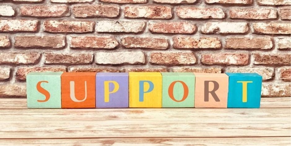 木製のカラフルなブロックで「SUPPORT」と並べられた文字、レンガの背景と木目の床