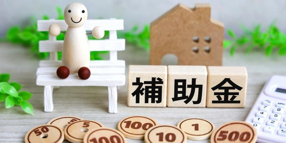 「補助金」の文字ブロックと家の模型、木製コインや電卓が並ぶ住宅支援イメージ